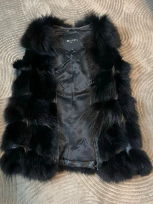 Svart pälsväst från Hollies - Säljer en fluffig svart pälsväst från Hollies med bred krage och lyxig känsla. Västen har ett glansigt innerfoder och är gjord av mjuk päls i blockmönster. Perfekt för dig som vill ha en statement piece till din outfit.