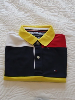 Färgglad piké från Tommy Hilfiger - Snygg pikétröja från Tommy Hilfiger i färgblock med gul krage, vita, röda och marinblå partier samt gul kant nertill. Klassisk pikéknäppning och liten logga på bröstet. Perfekt för dig som gillar preppy och färgstark stil.
