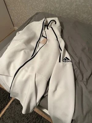 Vit hoodie från Adidas med svart detalj - Snygg vit hoodie från Adidas med svart dragkedja och svarta detaljer längs sömmarna. Adidas-logga på ärmen ger en sportig vibe. Hoodien har huva och kängurufickor framtill. Perfekt för dig som gillar streetwear och enkel stil.