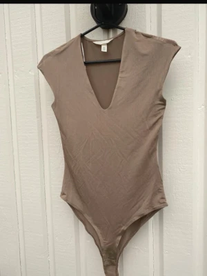 Beige body nyskick!  - Säljer en stilren beige body från H&M med djup v-ringning och korta ärmar. Lätt skimmrigt material, använd 1 gång bara. Säljes pga passar ej längre. Perfekt att matcha med jeans eller kjol för en clean look! Djur och rökfritt hem 