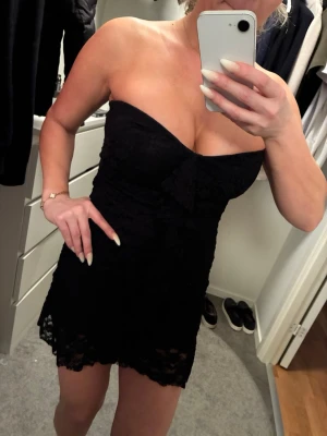 Svart axelbandslös spetsklänning - Säljer en svart, kort axelbandslös klänning i spets med volangdetaljer. Klänningen har en figurnära passform och är perfekt för dig som vill ha en elegant och stilren look. Spetsen ger en romantisk känsla och den korta längden gör den extra trendig.