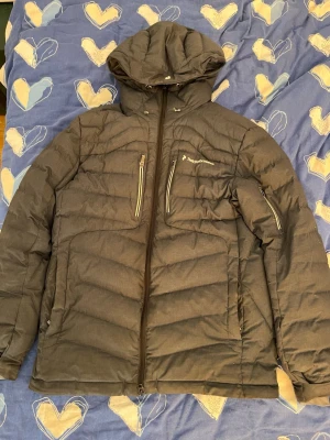 Svart dunjacka från Peak Performance - Snygg svart dunjacka från Peak Performance med huva och flera dragkedjeförsedda fickor. Jackan har quiltad design och är perfekt för kalla vinterdagar. Klassisk look med logga på bröstet och praktiska detaljer som hög krage och justerbar huva.