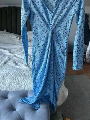 Blommig blå långklänning från Gina Tricot - Supersöt långklänning i ljusblått från Gina Tricot med svart blommigt mönster. Klänningen har v-ringning, krage och långa ärmar. Materialet är lätt och känns skönt mot huden. Perfekt för dig som gillar en feminin och trendig stil.