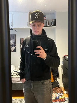 Svart Nike zip-hoodie med huva - Säljer en svart Nike hoodie med dragkedja och huva. Hoodien har vita detaljer längs bröstet och Nike-logga på vänster sida. Praktiska fickor framtill och justerbara snören i huvan. Perfekt för dig som gillar sportig och avslappnad stil.