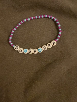 Färgglatt pärlarmband med text - Ett handgjort armband med lila och ljusblå små pärlor samt vita bokstavspärlor som stavar 'DU ÄR BRA' med en blå stjärna och ett blått hjärta. Armbandet har en lekfull och personlig stil, perfekt för att ge bort till någon speciell.