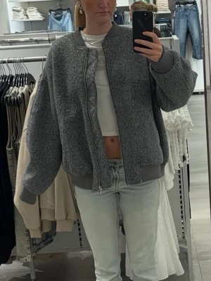 Grå teddyjacka med dragkedja - Mysig grå teddyjacka med oversized passform och ribbade muddar vid ärmslut och nederkant. Jackan har en hög krage och stängs med dragkedja framtill. Perfekt för att hålla sig varm och stilren under kyliga dagar.