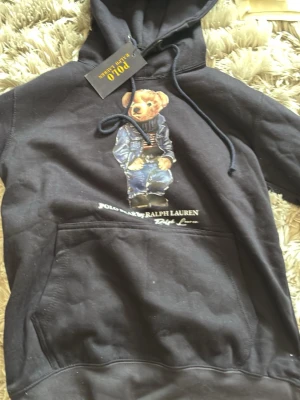 Svart hoodie Polo Ralph Lauren - Svart hoodie från Polo Ralph Lauren med ikoniska björnmotivet på bröstet. Stor känguruficka framtill och justerbar huva med snören. Perfekt för en avslappnad och trendig stil.
