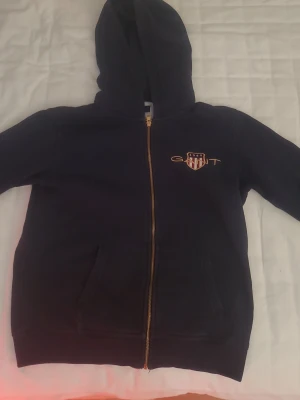 Navy blue zip hoodie från Gant - Mörkblå zip hoodie från Gant med guldfärgad dragkedja och broderad logga på bröstet. Klassisk huva och två fickor framtill. Tillverkad i mjukt sweatshirtmaterial som är skönt att ha på sig. Den är passad för 170cm lång människa.