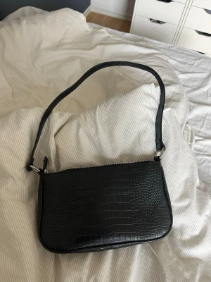 Svart axelväska i croco-mönster - Stilren svart axelväska med croco-mönstrad yta och silverfärgad dragkedja. Väskan har en avlång, mjukt rundad form och justerbar axelrem. Invändigt finns ett mindre fack med dragkedja. Perfekt för dig som gillar trendiga accessoarer.
