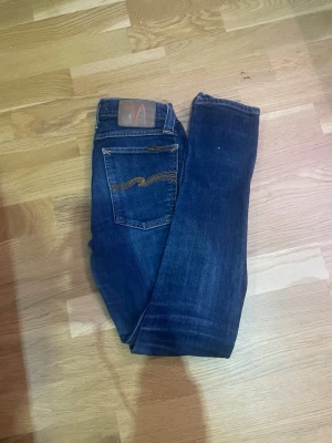 Mörkblå jeans från Nudie Jeans - Snygga mörkblå jeans från Nudie Jeans med klassisk femficksdesign och kontrastsömmar på bakfickan. Jeansen har en rak passform och är tillverkade i denim av bomull. Perfekta för en avslappnad och trendig look.