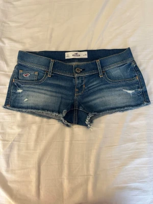 Låg midja blå jeansshorts från Hollister - Säljer ett par blå denimshorts från Hollister med låg midja och råa kanter. Shortsen har slitna detaljer, fem fickor och klassisk knappgylf. Perfekta för sommaren och ger en avslappnad look.