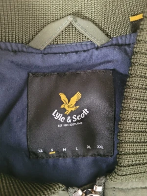 Olivgrön bomberjacka från Lyle & Scott - Snygg olivgrön bomberjacka från Lyle & Scott med ribbade muddar och krage. Jackan har dragkedja framtill och är tillverkad i ett lätt, slitstarkt material. Perfekt för dig som vill ha en stilren och klassisk look. Använd 1 gång.