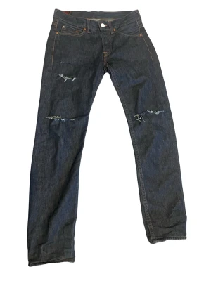 Mörkblå slitna jeans från Evisu - Säljer ett par mörkblå jeans från Evisu med slitningar och hål på knäna. Klassisk femficksmodell med orange sömmar och bruna detaljer på bakfickorna. Raka ben och tillverkade i kraftig denim. Perfekta för en streetwear-look.!OBS! Dessa på bilden är lite uppsydda så dm mig för exakt läng på dom !