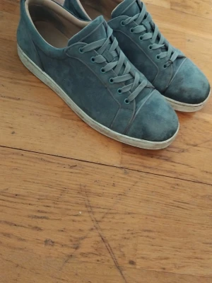 Blåa sneakers röd sula - Snygga blåa sneakers i mocka  med klassisk röd sula och vita kanter. Skorna har lågt skaft, rund tå och matchande blå skosnören. Perfekta för dig som vill sticka ut med exklusiva detaljer.