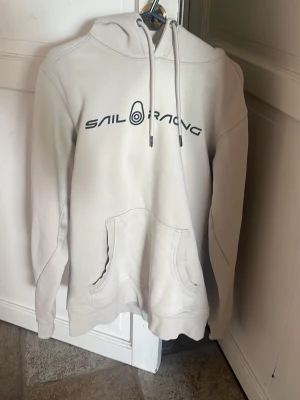 Vit hoodie från Sail Racing - Vit hoodie från Sail Racing med svart logga och tryck på bröstet. Klassisk känguruficka framtill. Säljer den för har vuxit hur den, det står S men passar M. Pris går diskutera!