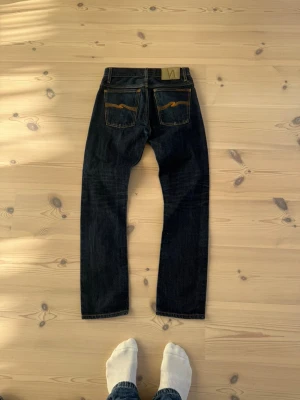 Mörkblå jeans från Nudie Jeans - Säljer ett par mörkblå jeans från Nudie Jeans med klassisk rak passform. Jeansen har kontrastsömmar, två bakfickor med Nudie-signatur och är tillverkade i kraftig denim. Perfekta för en avslappnad och stilren look.