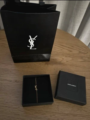 Guldfärgat armband från Saint Laurent - Snyggt och stilrent armband från Saint Laurent i guldfärgad metall med den ikoniska YSL-loggan som hänge. Tunt och elegant kedjearmband som passar perfekt till dig som gillar exklusiva accessoarer med minimalistisk design.