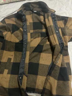 Rutig overshirt i brunt och svart - Overshirt med stora bruna och svarta rutor, klassisk skjortkrage och tryckknappar framtill. Två stora bröstfickor med lock och snedställda sidfickor. Tjockt tyg som ger en robust känsla, perfekt för lager-på-lager-stil.