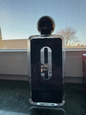 Afnan 9pm Eau de Parfum 100ml - Afnan 9pm Eau de Parfum i en stilren, rektangulär glasflaska med svart etikett och stor silverfärgad '9pm'-logga. Flaskan har en rund, svart kork och rymmer 100 ml. Doften är känd för sin intensiva och moderna karaktär.