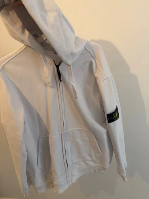Vit zip hoodie från Stone Island - Säljer en vit hoodie från Stone Island med dragkedja framtill och klassisk logopatch på vänster ärm. Hoodien har huva med dragsko och två fickor fram. Tillverkad i mjuk bomull, perfekt för en avslappnad streetwear-look. Storlek Xl passar L längd 183-186