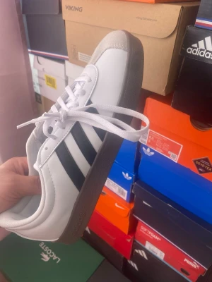 Adidas vita sneakers med svarta ränder - Klassiska Adidas sneakers i vitt skinn med de ikoniska tre svarta ränderna på sidan. Skon har vit ovandel, grå tåhätta i mocka och brun sula. Lågt skaft och vita skosnören. Perfekt för dig som gillar stilrena och tidlösa sneakers.