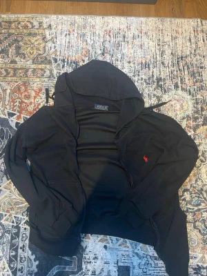 Svart hoodie från Polo Ralph Lauren - Svart hoodie med dragkedja från Polo Ralph Lauren. Klassisk design med huva och fickor framtill. Ikoniska röda Polo-loggan broderad på bröstet. Mjuk bomullskänsla och ribbade muddar vid ärmslut och nederkant. Perfekt för en avslappnad och stilren look.