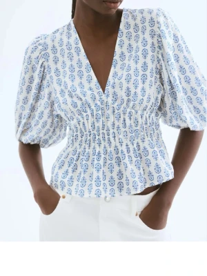 Vit blus med blått mönster och puffärm - Superfin vit blus med blått paisleymönster och djup V-ringning. Blusen har puffiga korta ärmar och smockad midja som ger en snygg siluett. Perfekt till jeans eller kjol för en fräsch look. Materialet känns lätt och luftigt.