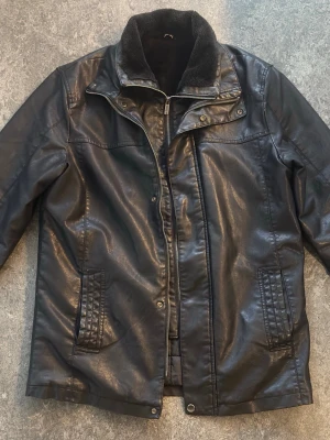 Svart skinnjacka med krage - Snygg svart skinnjacka med klassisk krage och dragkedja framtill. Jackan har två fickor med detaljerade sömmar och en mjuk, svart foderkrage. Perfekt för dig som vill ha en stilren och tidlös look.