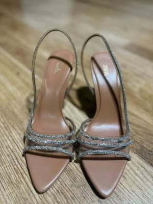 Beige pumps med strass från Zara - Säljer ett par snygga beige pumps från Zara med transparenta remmar dekorerade med glittrande strass. Skorna har spetsig tå, öppen häl och smal klack för en elegant look. Perfekta för att lyfta vilken outfit som helst.