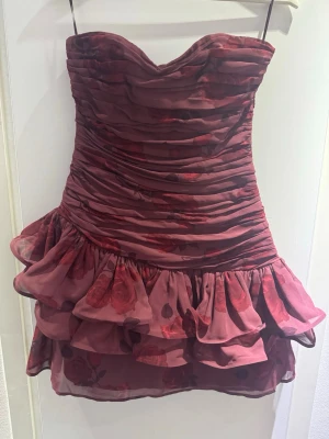 Vinröd miniklänning från Adoore - Supersnygg vinröd miniklänning från Adoore med volanger och draperad överdel. Axelbandslös modell med hjärtformad urringning och blommigt mönster. Perfekt för dig som vill sticka ut med en romantisk och trendig look.