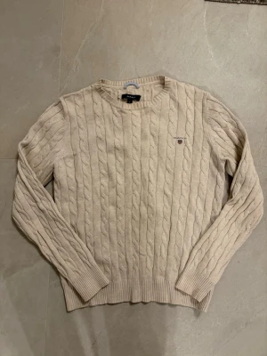Beige kabelstickad tröja från Gant - Kabelstickad tröja i beige från Gant med rund halsringning och diskret broderad logga på bröstet. Tröjan har långa ärmar och ribbade muddar vid ärmslut och nederkant. Perfekt för en stilren och klassisk look.