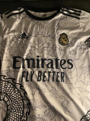 Real Madrid Adidas drakmönstrad tröja - Snygg vit Real Madrid-tröja från Adidas med svart drakmönster och svarta detaljer. Tröjan har klubbmärke, Adidas-logga och Emirates Fly Better-tryck på bröstet. Perfekt för fotboll eller streetwear.