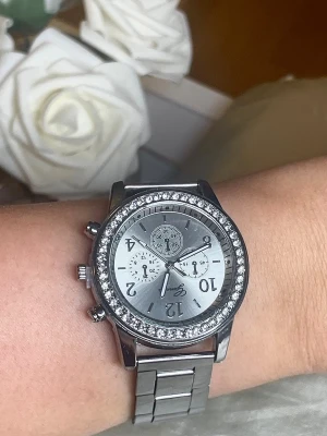 Silverfärgad klocka med stenar - Stilren klocka från Geneva i silverfärg med metallarmband och gnistrande stenar runt urtavlan. Urtavlan har flera dekorativa små visare och tydliga siffror. 