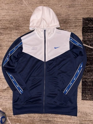 Nike Repeat hoodie - Nike hoodie i blått och vitt med hel dragkedja och huva. Framsidan har Nike-logga och Swoosh i blått. Ärmarna har blå och vita detaljer med Swoosh-mönster. Tillverkad i ett mjukt, sportigt material som känns skönt mot huden.