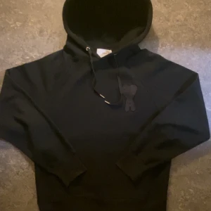 Svart hoodie från Ami Paris - Säljer en svart hoodie från Ami Paris med broderad logga i form av ett hjärta och bokstaven A på bröstet. Hoodien har dragsko i huvan och ribbade muddar vid ärmslut och nederkant. Perfekt för en stilren och trendig look. Passar även M