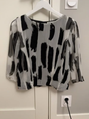 Svartvit mönstrad blus från H&M - Trendig blus från H&M i svartvitt abstrakt mönster. Tillverkad i ett lätt, transparent tyg med trekvartsärm och rund halsringning. Perfekt för dig som gillar en modern och edgy stil.