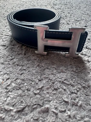 Marinblått skärp med H-spänne - Snyggt matinblått bälte från H med ett stort, silverfärgat H-spänne. Bältet har vita kontrastsömmar och är gjort i äkta skinn. Perfekt accessoar för att lyfta vilken outfit som helst.