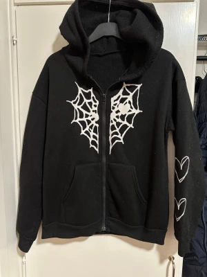 Svart hoodie med spindelnät och hjärtan strl: S - Svart hoodie med dragkedja och huva. På bröstet finns vita spindelnätsmotiv och på ena ärmen två vita hjärtan. Hoodien har känguruficka framtill och ribbade muddar. Perfekt för dig som gillar alternativ stil och coola detaljer. Noppor förekommer. 
