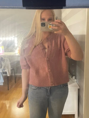 Kort rosa kofta med knappar - Säljer en kort kofta i mjukt rosa material med bred ribbstickad kant och stora matchande knappar framtill. Koftan har korta ärmar och en djup v-ringning, perfekt att styla med jeans eller kjol.