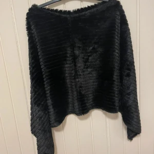 Svart ribbad fuskpäls-sjal - Mysig svart sjal i fuskpäls med ribbad struktur. Sjalens form är asymmetrisk och materialet är mjukt och glansigt, perfekt för att ge en lyxig touch till din outfit. Enkel att slänga över axlarna för en trendig look.