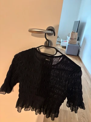 Svart croppad topp från Gina Tricot - Trendig svart croppad topp från Gina Tricot med transparent, rynkad mesh och volangkant nertill och vid ärmslut. Perfekt för att lyfta din outfit med en edgy vibe. Kortärmad och figurnära passform.