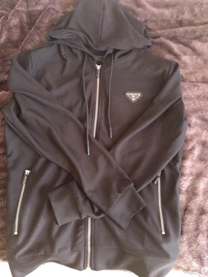 Svart hoodie med dragkedja från Prada - Snygg svart hoodie från Prada med dragkedja framtill och två fickor med dragkedjor. Klassisk huva med snören och ikonisk Prada-logga på bröstet. Tillverkad i mjukt material som känns lyxigt och bekvämt. Perfekt för dig som gillar exklusiv streetwear. Pris kan diskuteras