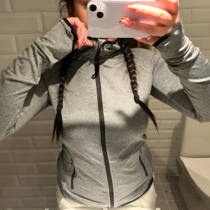 Grå sportig hoodie med dragkedja - Snygg grå hoodie med hel dragkedja och svarta detaljer. Tröjan har huva, två fickor med dragkedja och är tillverkad i ett stretchigt funktionsmaterial. Perfekt för träning eller chill dagar. Tight passform och modern look.