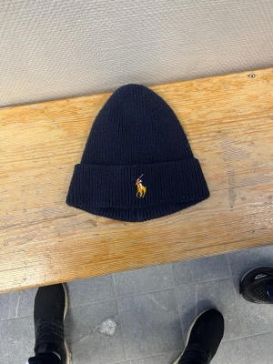 Mörkblå stickad mössa från Polo Ralph Lauren - Snygg mörkblå stickad mössa från Polo Ralph Lauren med klassisk broderad logga framtill i gult och rött. Mössan har uppvikt kant och är tillverkad i mjukt, ribbstickat material som håller dig varm under kalla dagar. Sitter riktigt bra 😊 