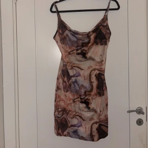 Mönstrad kort klänning med smala axelband - Säljer en kort klänning med smala axelband och ett unikt mönster i bruna, beige och lila toner. Klänningen har en figurnära passform och är perfekt för dig som gillar trendiga och färgstarka plagg. Materialet känns mjukt och följsamt mot kroppen.
