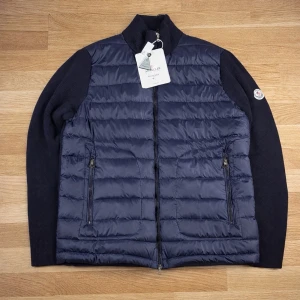 Moncler Cardigan - Snygg blå Cardigan från Moncler med quiltad front och rygg i glansigt material samt stickade svarta ärmar. Jackan har två dragkedjefickor fram och hög krage. Ikonisk Moncler-logga på vänster ärm. Perfekt för kalla vinterdagar. TVEKA INTE ATT SKICKA EN BUD:)