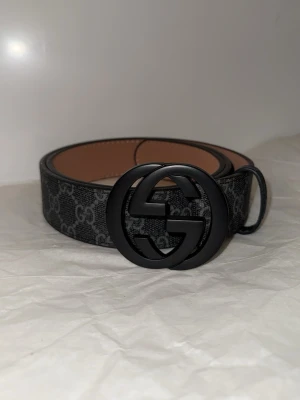 Svart Gucci skärp med GG-spänne - Snyggt svart skärp från Gucci med klassiskt GG-mönster och stort svart GG-spänne. Bältet är gjort i skinn med detaljer i mörkgrått och svart, och insidan är i ljust läder. Perfekt accessoar för att lyfta vilken outfit som helst.