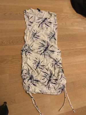 Vit och blå kort klänning med snörning - Snygg vit kort klänning med blått mönster och snörning i sidorna. Klänningen har ärmlös design och ett coolt abstrakt mönster som ger en unik look. Perfekt för varma dagar och sommarens alla äventyr.