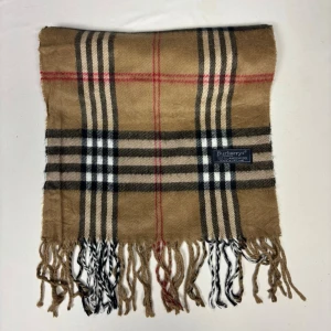 Beige Vintage rutig halsduk från Burberrys  - Klassisk beige vintage halsduk från Burberrys med det ikoniska rutmönstret i svart, vitt och rött. Halsduken har fransar i ändarna och är tillverkad i mjuk ull. Perfekt accessoar för att lyfta din outfit med en tidlös och lyxig känsla.