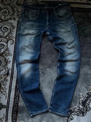 Blå Replay Anbass jeans - Säljer ett par blå jeans från Replay, modell Anbass. Jeansen har en snygg tvätt med slitningar och klassisk femficksdesign. De är raka i benen och har en modern passform som funkar till det mesta. Perfekta för dig som gillar en avslappnad men stilren look.